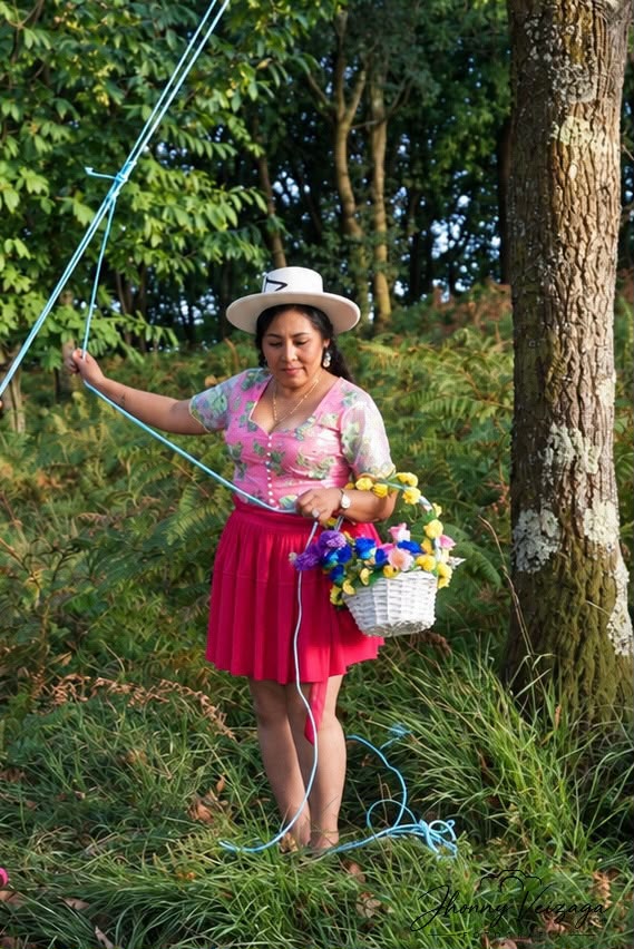 Una cholita con sombrero blanco y pollera fucsia sostiene una canasta de flores junto a un árbol